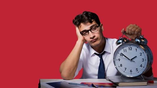 A young man sits at his desk, visibly exhausted, with one hand on a giant alarm clock.