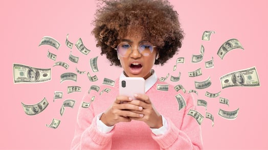 A young Black woman in a pink sweater and glasses looks at her phone in disbelief as money flies out.
