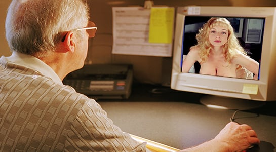 A man sits at a computer looking at a monitor displaying a woman in lingerie.