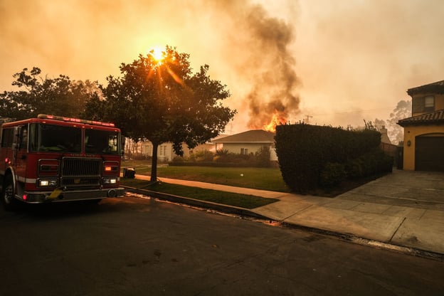 A red fire truck is parked on a street with a house engulfed in flames and smoke in the background, under an orange sky.