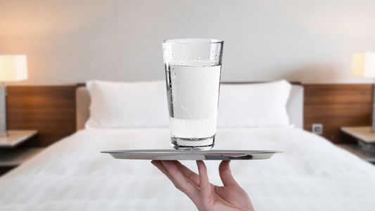 A hand holding a silver tray with a glass of water on it in front of a hotel bed. 