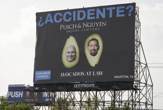 A black roadside billboard shows two lawyers' faces inside two halves of an avocado with the text: "¿ACCIDENTE? PUSCH &amp; NGUYEN INJURY LAWYERS AVOCADOS AT LAW".