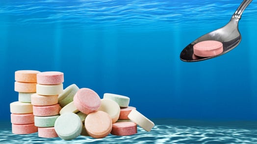 A giant metal spoon sinks a pink Tums tablet into the ocean, where several other tablets already sit on the floor.