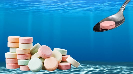 A giant metal spoon sinks a pink Tums tablet into the ocean, where several other tablets already sit on the floor.
