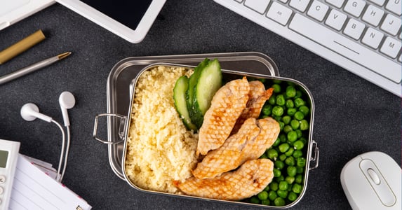  A metal container with peas, chicken, cucumbers, and rice sits on an office desk. Also on the desk are a keyboard, mouse, and earbuds. 