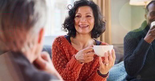 A smiling woman in a sweater holding a cup of tea sits chats with two men.