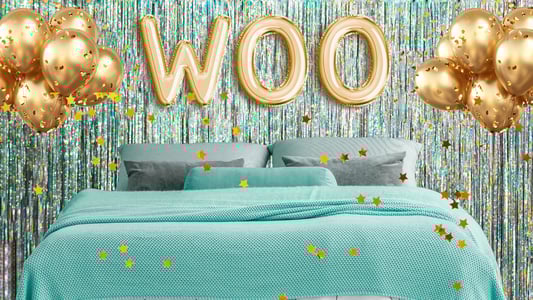 A bed topped with a teal bedspread and gray pillows surrounded by gold balloons, including three gold balloons that spell out, 