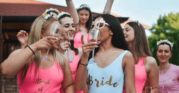 A young woman n a swimsuit that reads 'bride' toasts another woman in a swimsuit that reads 'maid of honor,' while several other bridesmaids stand in the background.