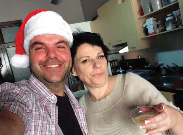 A selfie-style photo of a smiling Johnathan Walton wearing a Santa hat next to a woman, Mair Smyth, holding a small glass in a kitchen setting.